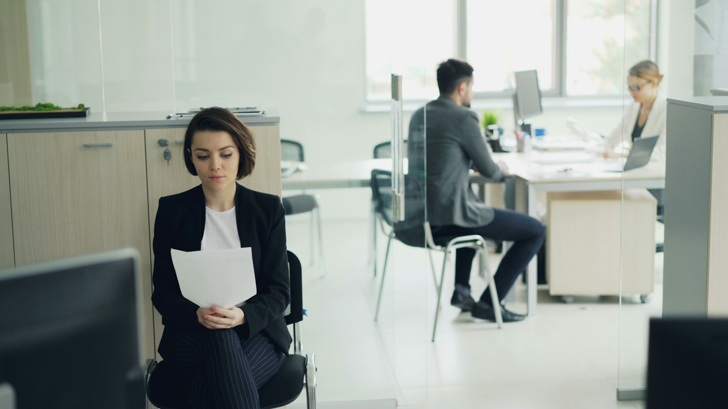 Woman in office holding papers, colleagues work behind glass.